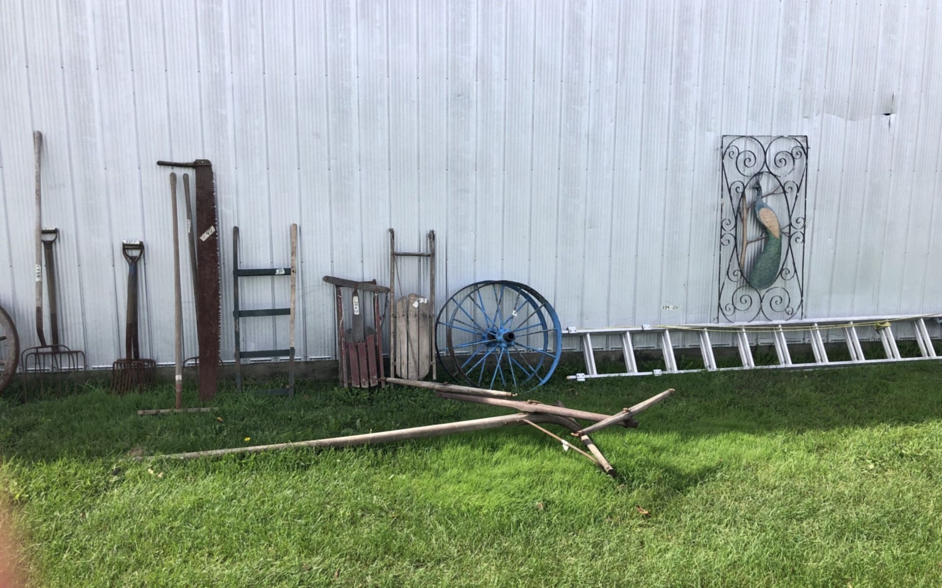 Old hand tools, a ladder, a wagon wheel and an iron panel against a steel building (photograph from the JMAC Auctions website)